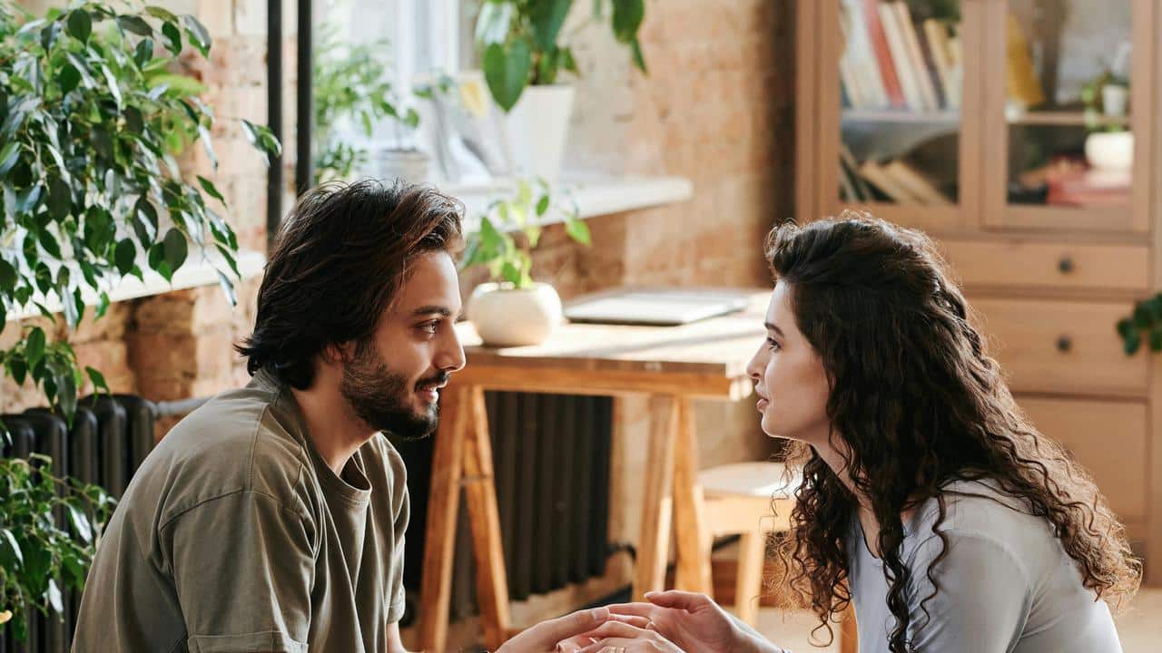 A couple sitting indoors holding hands and talking.