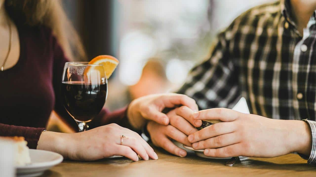A couple holding hands at a table beside a glass of wine.