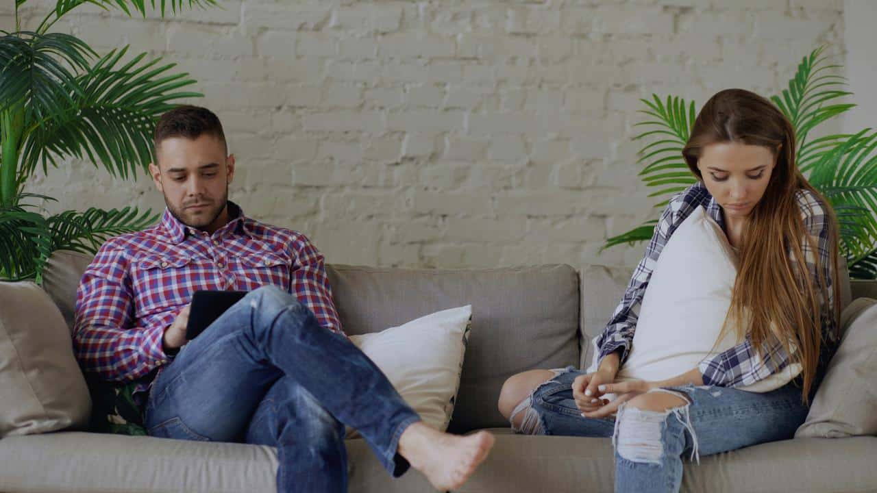 A man and a woman sitting apart on a couch, both looking down and disengaged.
