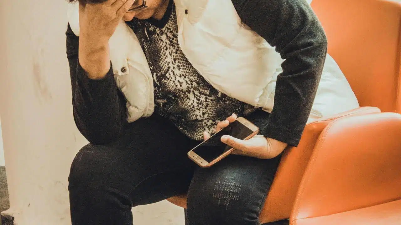 A worried person sitting on a chair holding a smartphone and covering their face.