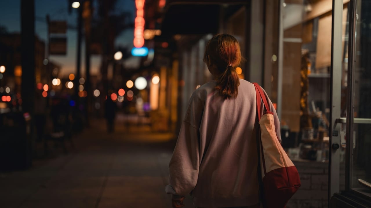 Back view of a person with a ponytail walking down a city sidewalk at night.