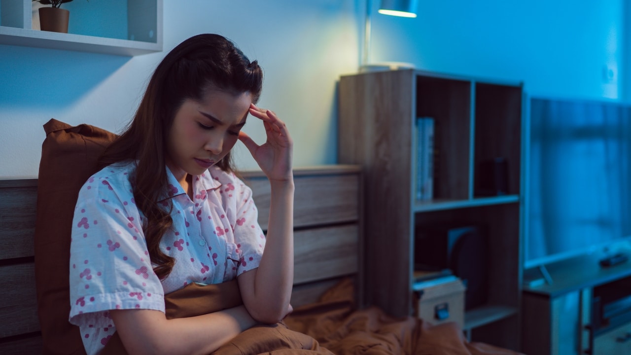 A woman in patterned pajamas sits on a bed looking down with a hand to her forehead.