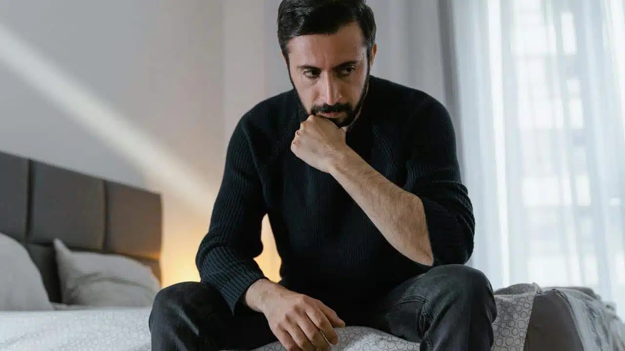 A man in a black sweater sitting on a bed with a pensive expression.