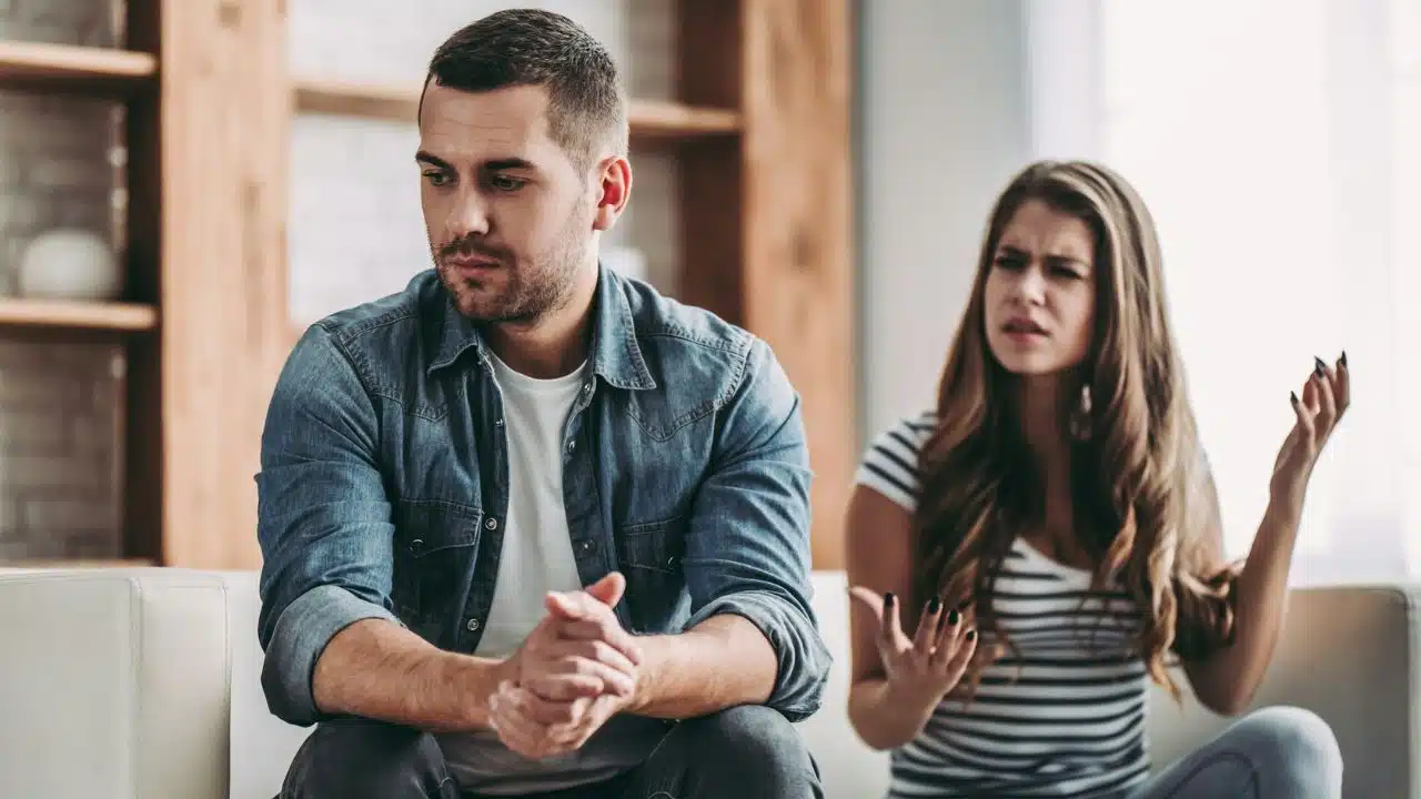 A man looks away while a woman behind him gestures and speaks with a frown.