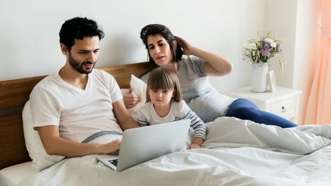 A man uses a laptop in bed while a woman and a child sit nearby.