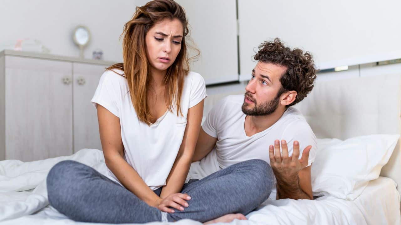 A woman sits cross-legged on a bed while a man gestures with one hand toward her.