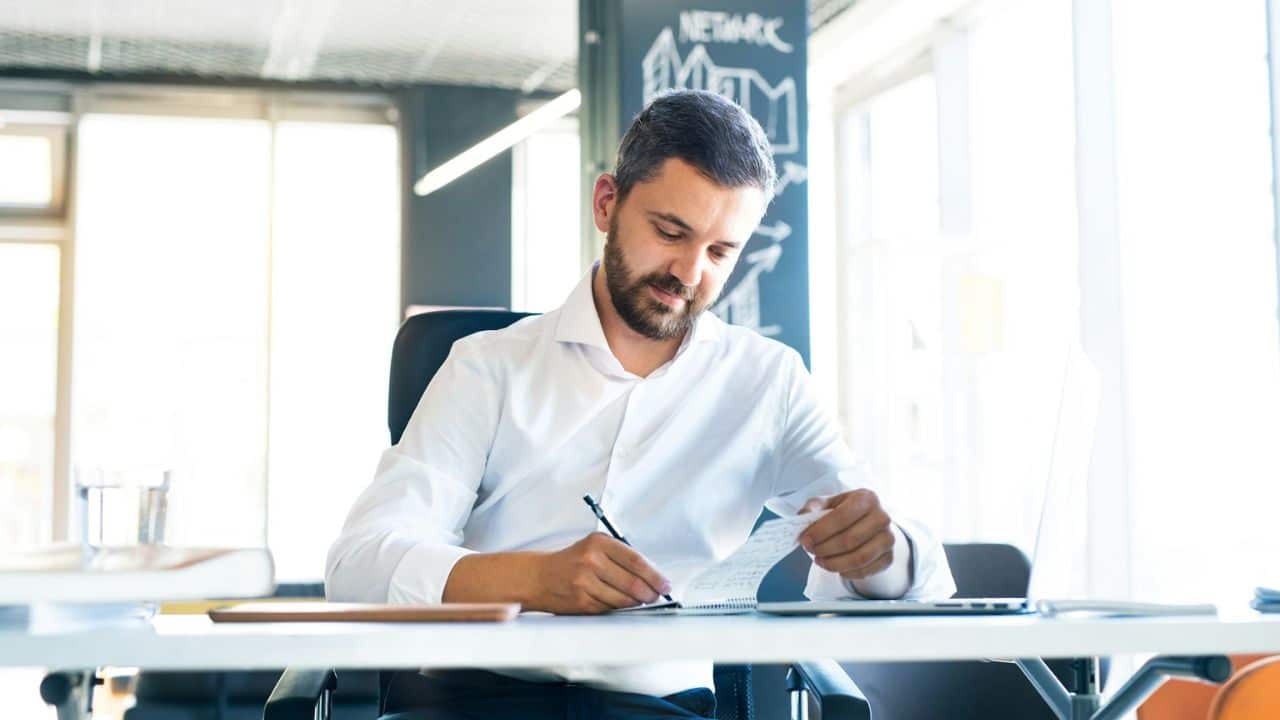 A man with a beard and dark hair sits at a desk, writing in a notebook.