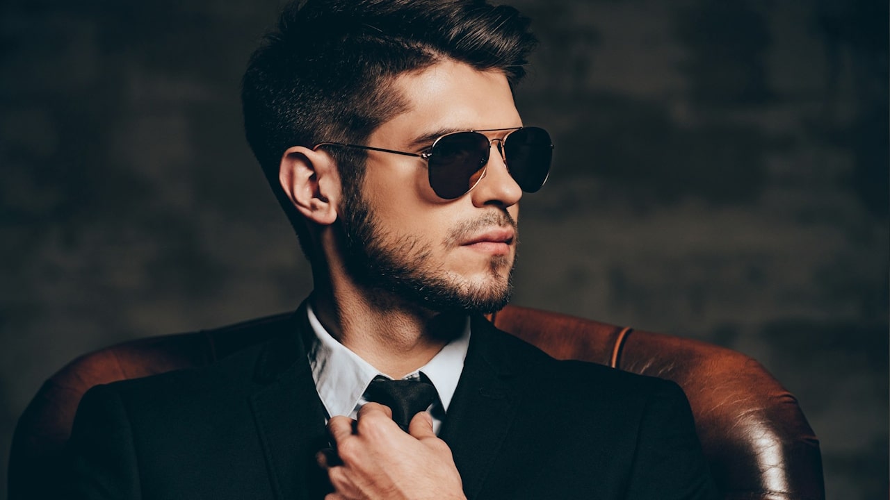 A stylish man in a black suit and aviator sunglasses sitting in a leather chair, looking away while adjusting his black tie.