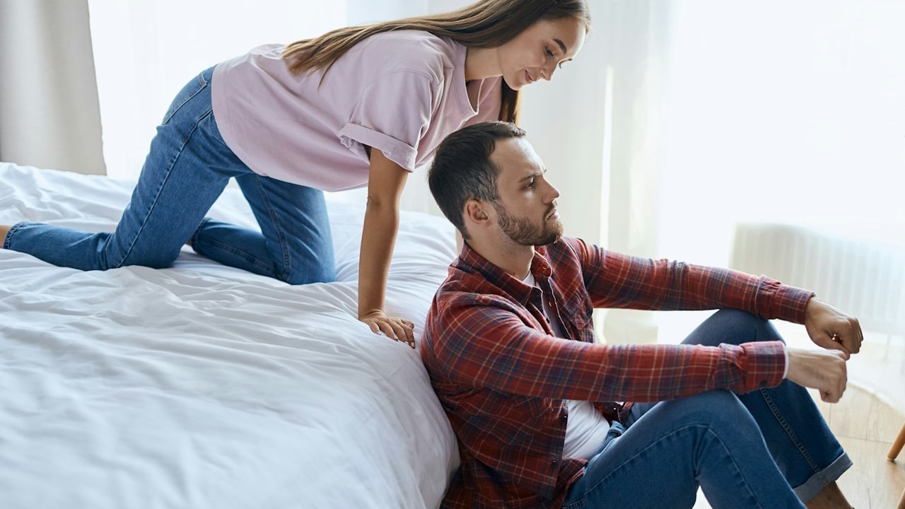 A woman on all fours on a bed looking at her husband, who is looking a bit angry.