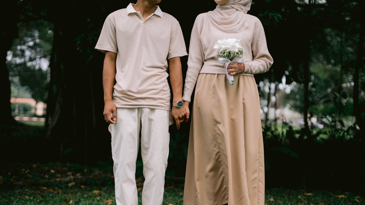 A couple standing on grass in matching beige/off-white attire, holding hands, with the woman holding a small white bouquet.