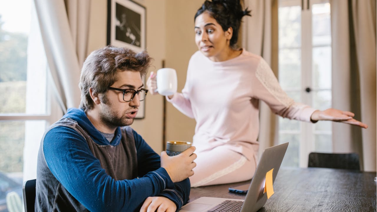 A man working on a laptop is interrupted by an angry woman holding a mug