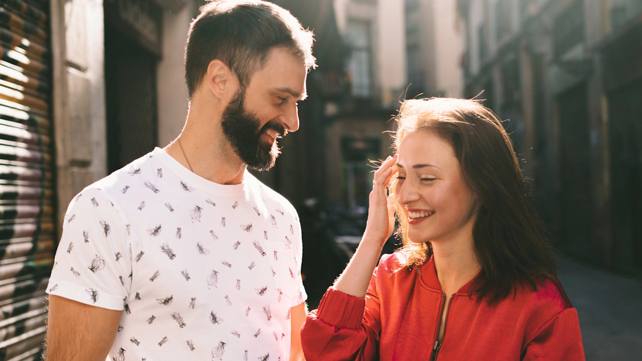 A smiling couple on a sunny street, the woman touching her hair.