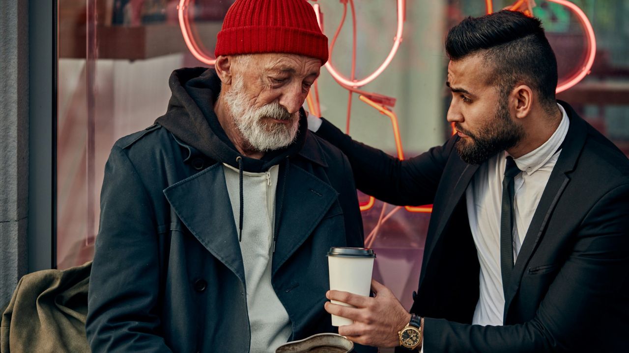 A younger man in a suit is offering a cup of coffee to an older man with a beard wearing a red knit hat and coat.