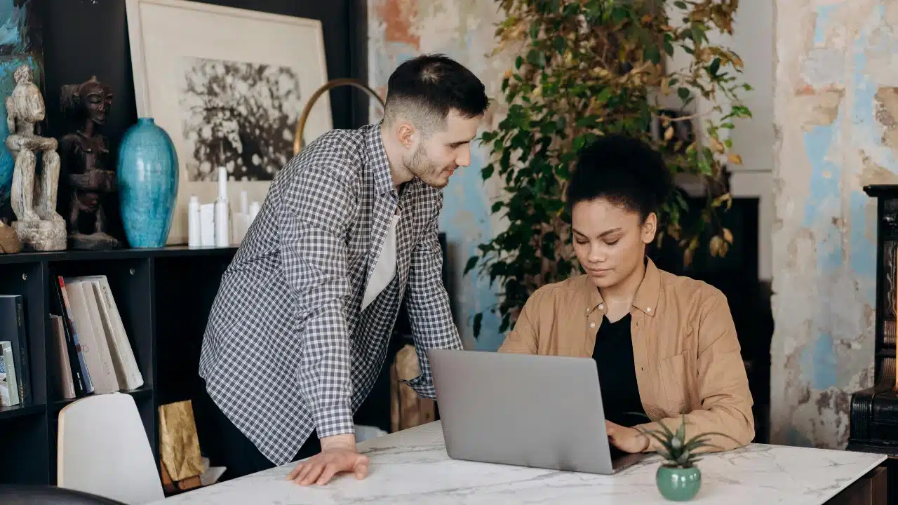 A man in a plaid shirt stands beside a woman who is typing on a laptop.
