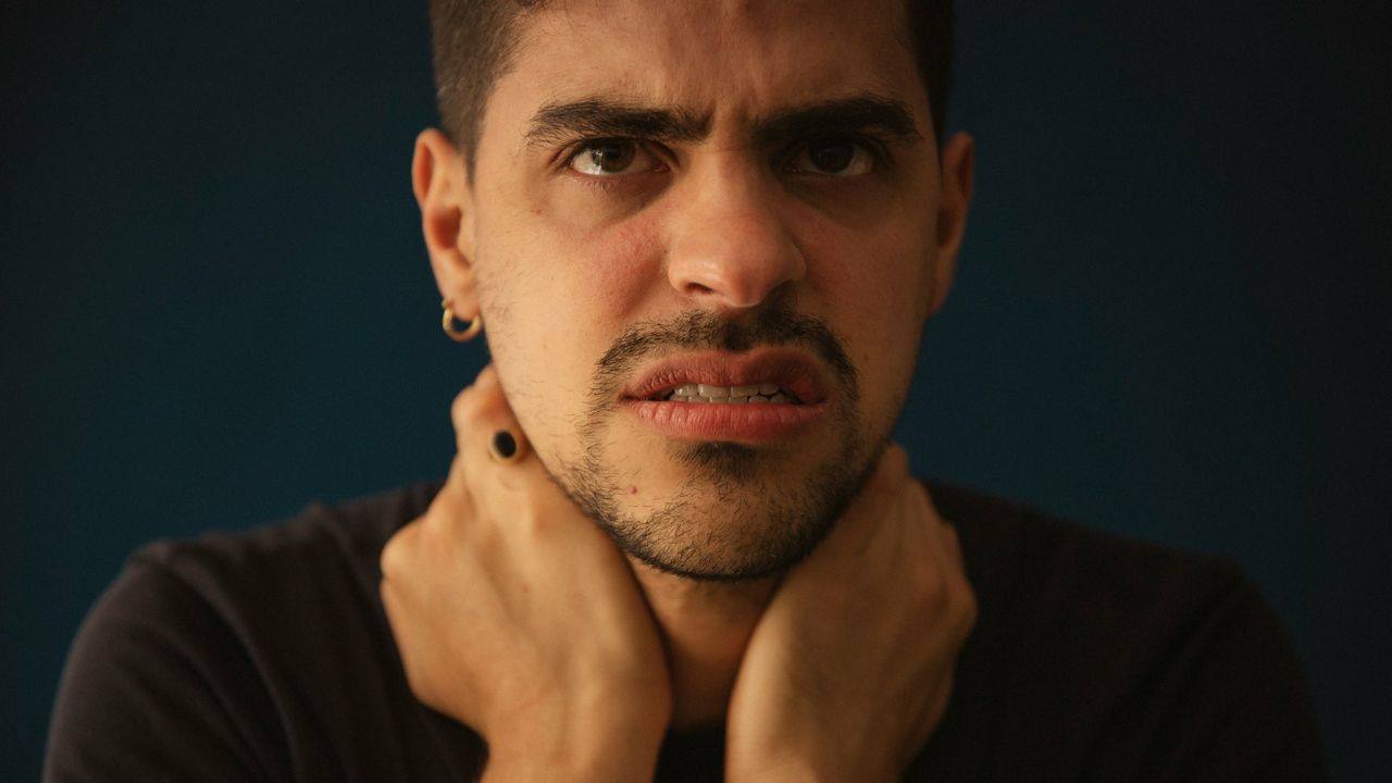 Close-up of a man with a beard and earrings looking angry or distressed, clutching his neck.