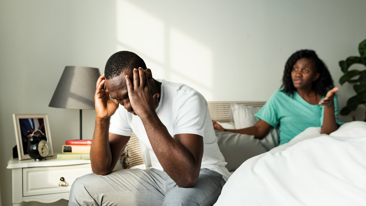 A man holding his head in his hands and looking stressed while sitting on a bed with a woman in the background who’s telling him something.