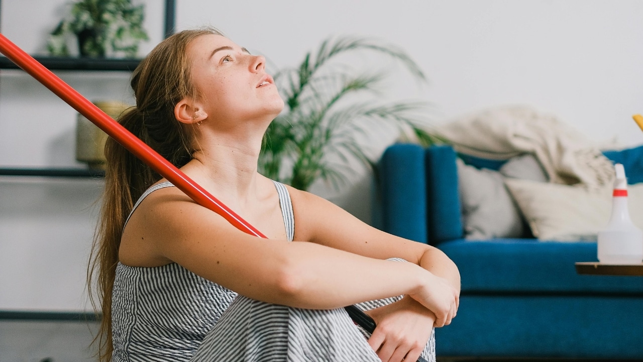 A woman sits on the floor holding a red mop handle while looking upward thoughtfully.