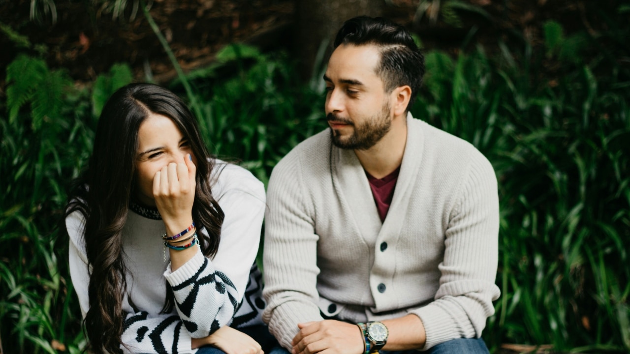 A man in a cardigan looks at a woman who is laughing and covering her mouth.
