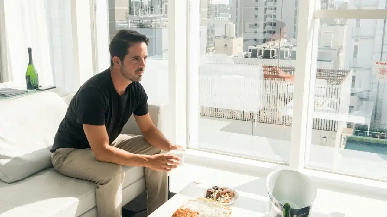 A man in a black shirt sits on a white sofa, looking out a window.