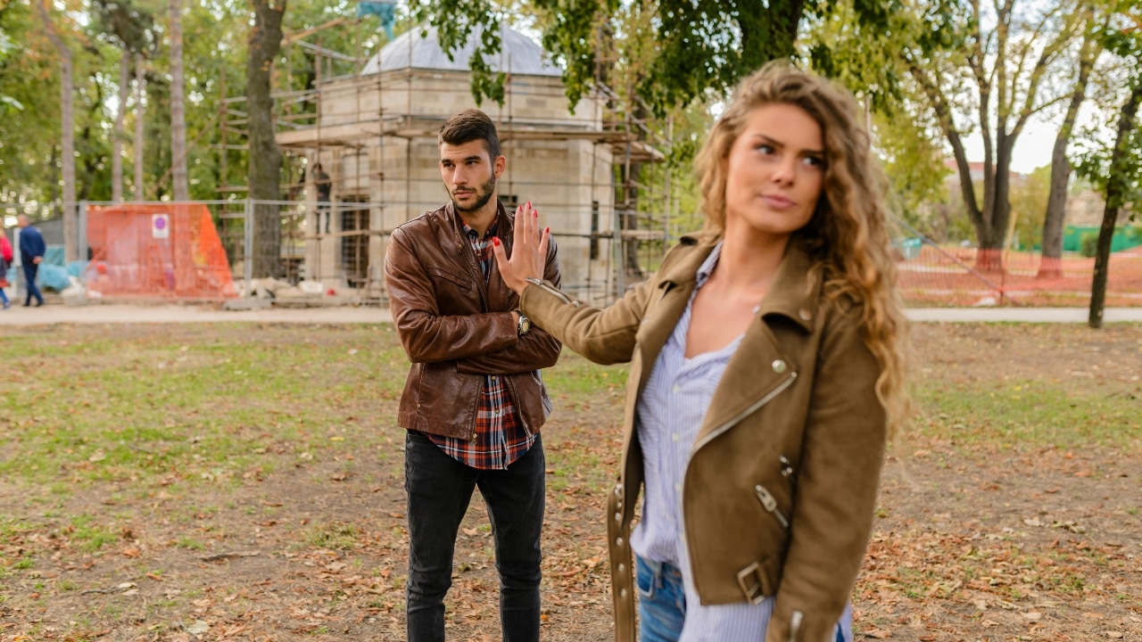 A woman extends her hand in a stop gesture toward a man standing in a park.