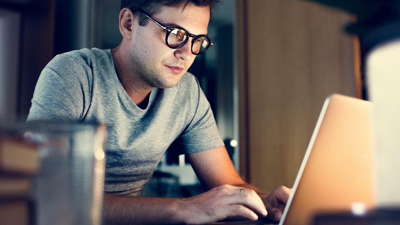 A man wearing glasses looks intently at a glowing laptop screen in a dimly lit room.
