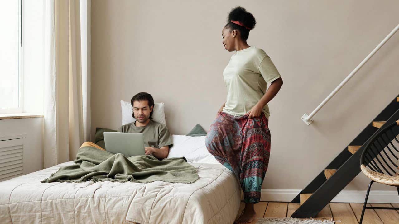 A woman stands near a bed where a man sits under blankets using a laptop.
