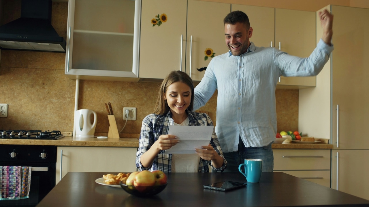 A woman reads a paper while a man stands behind her with raised arms.