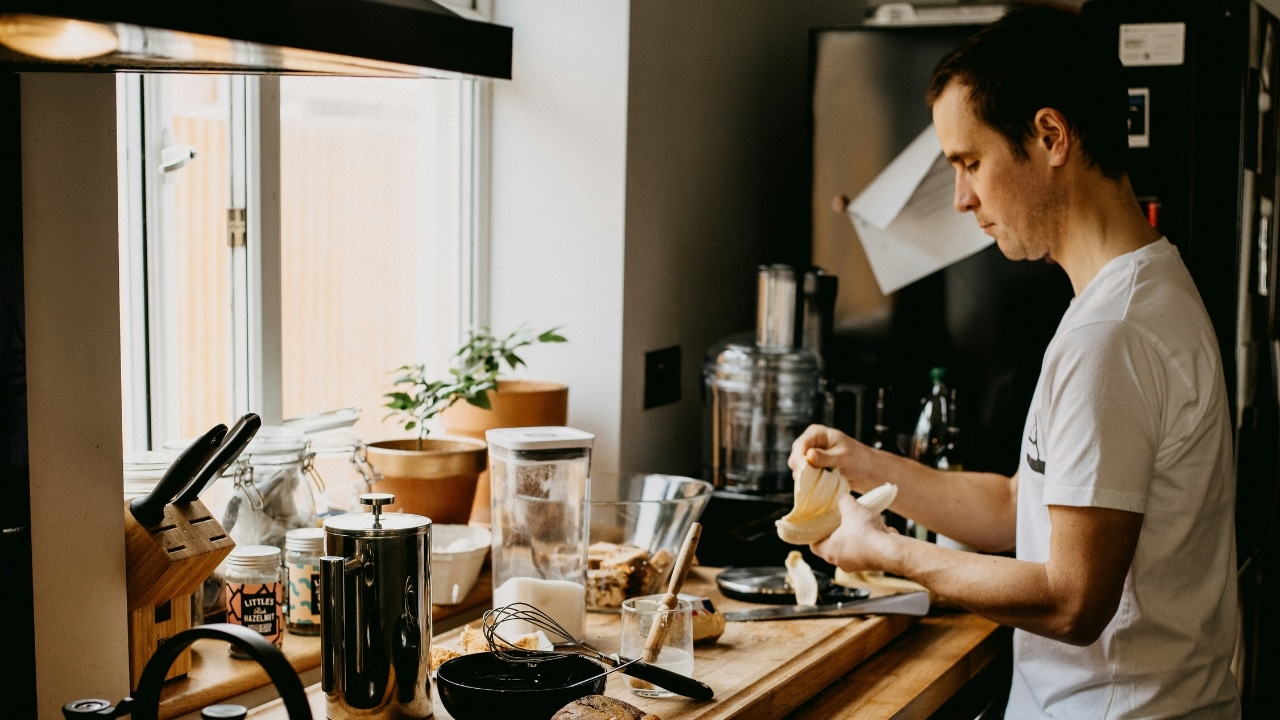 A man in a white shirt peels a banana in a kitchen filled with appliances.