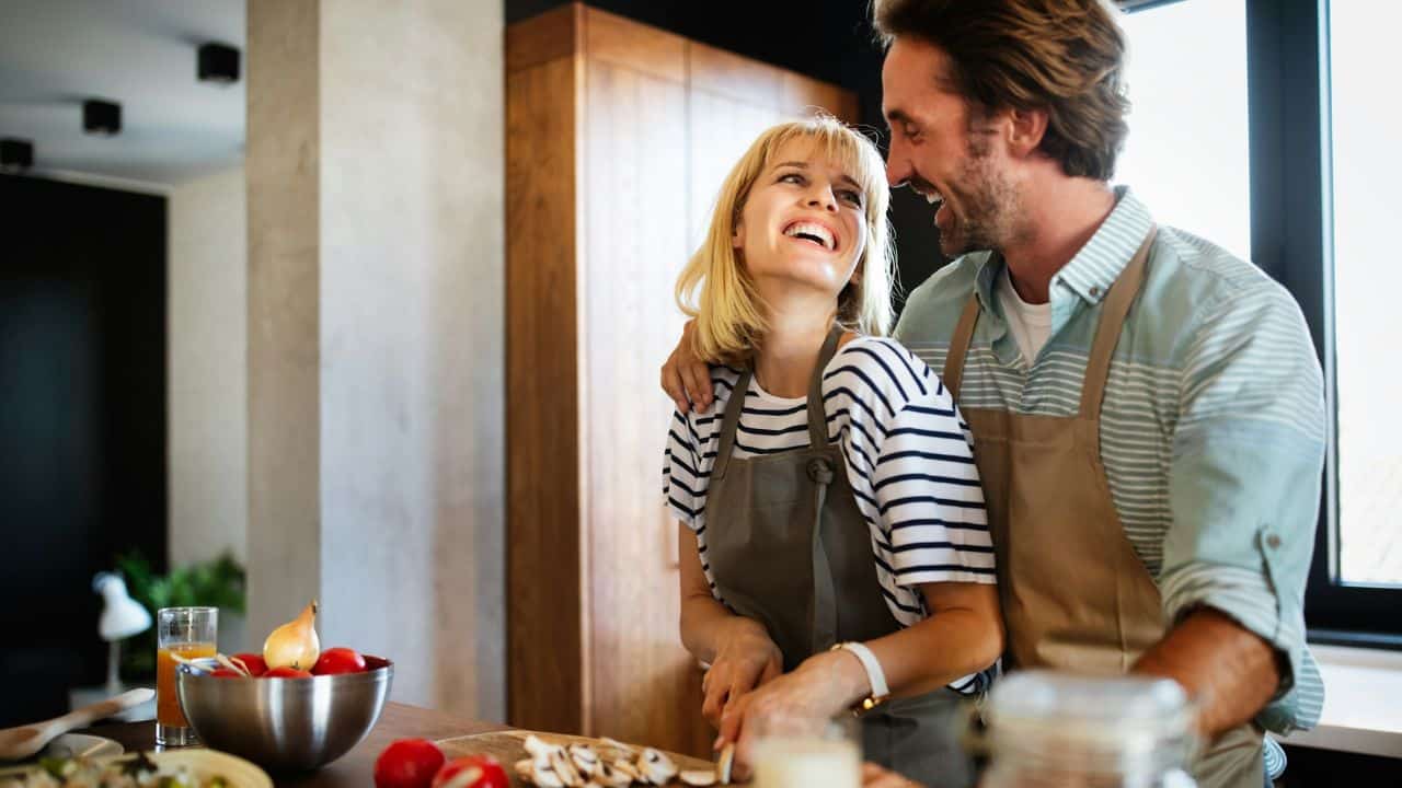 A man and woman in aprons laugh together while preparing food in a bright kitchen.