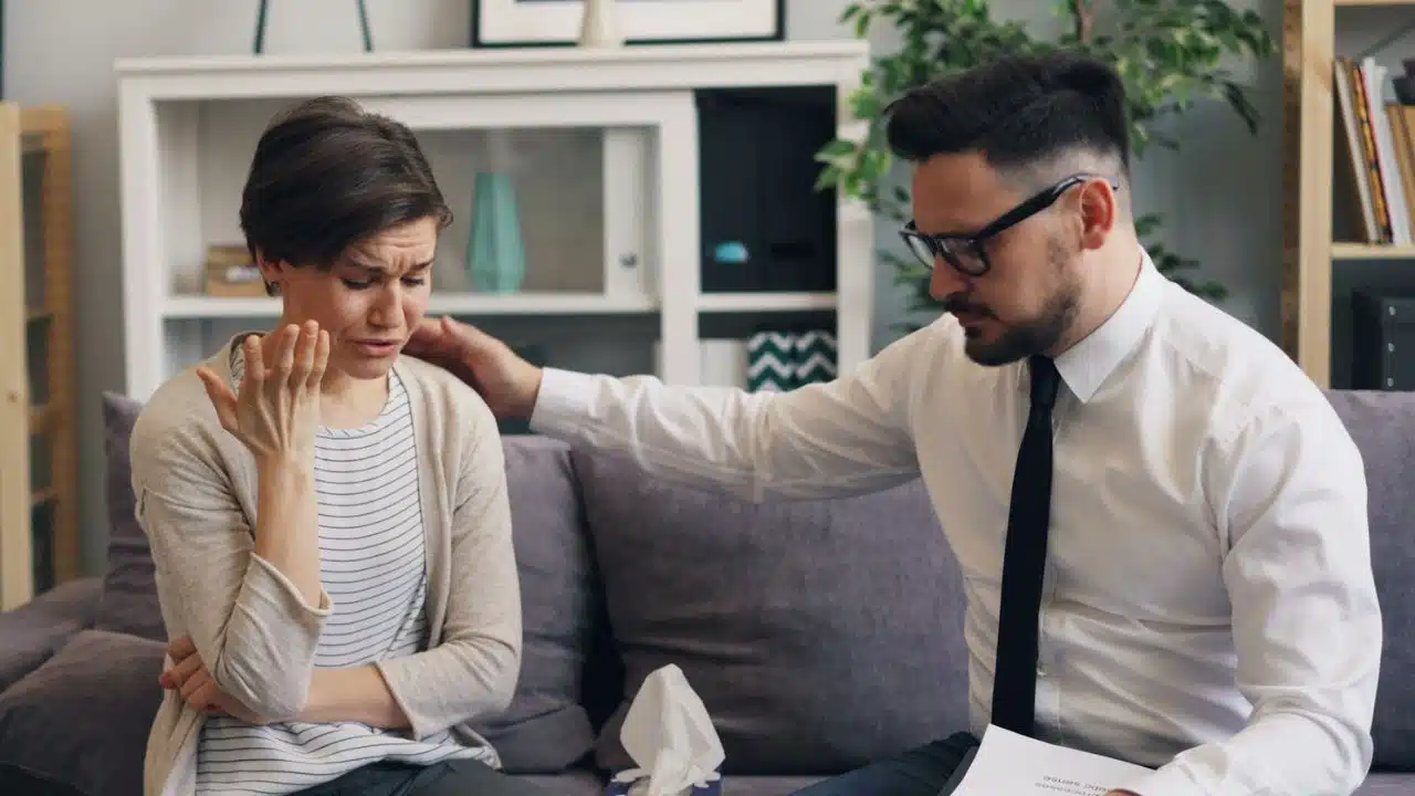 A woman cries on a couch while a man in a white shirt comforts her shoulder.