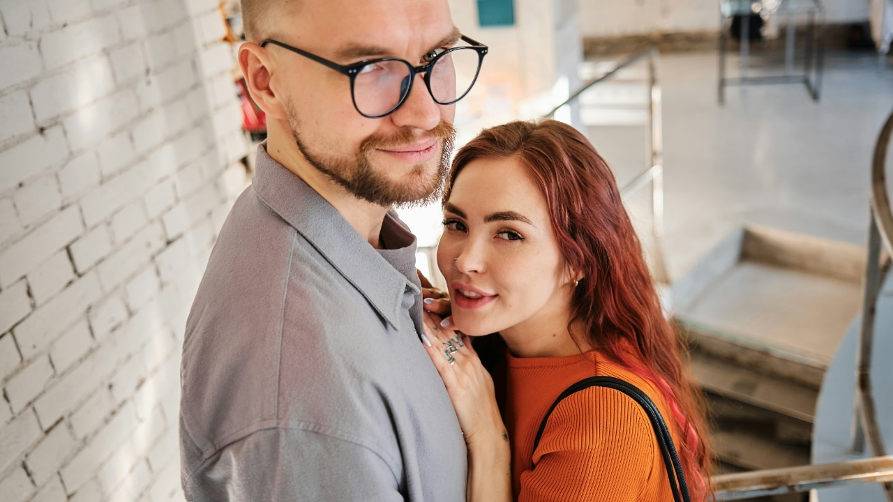 A woman with red hair leans against a man’s chest while they both look at camera.