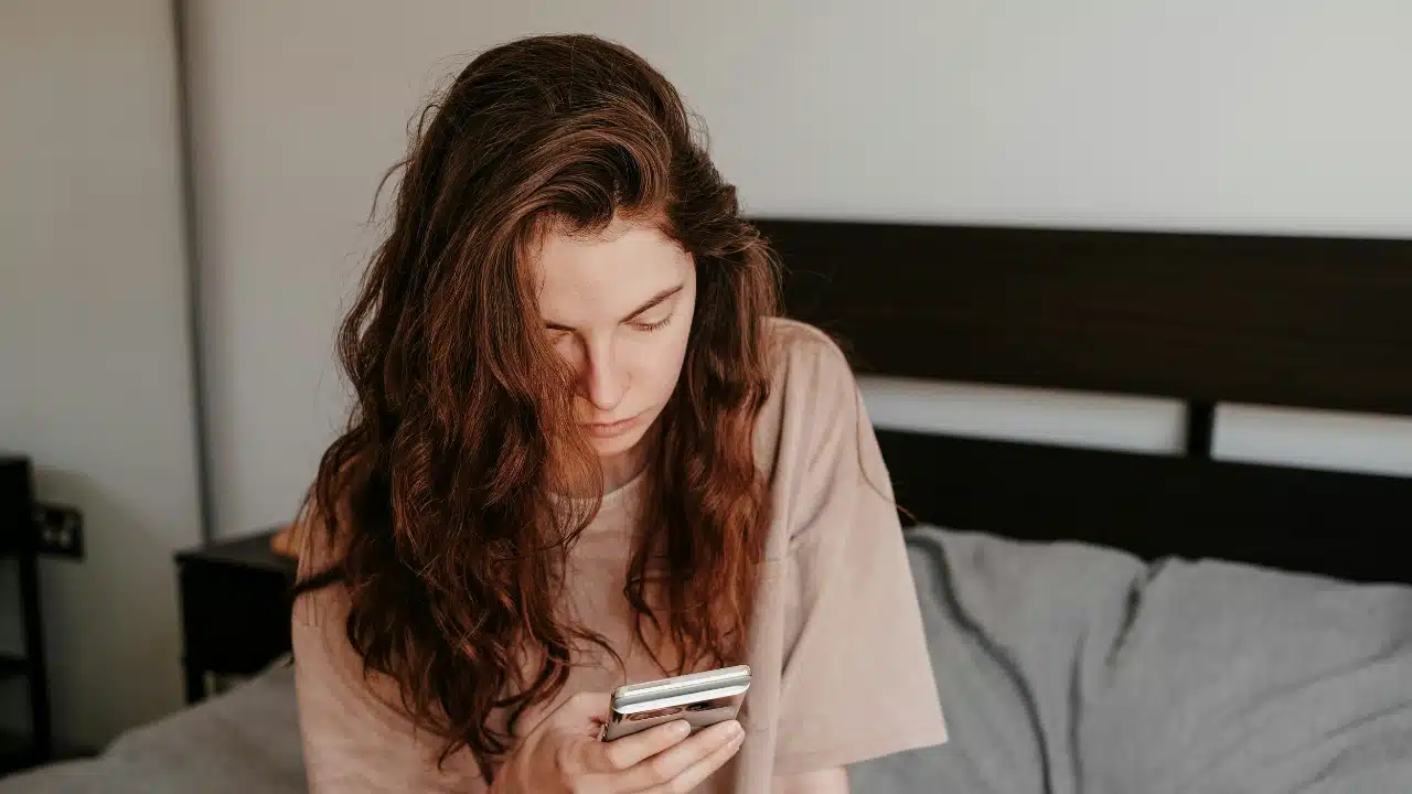 A woman with long wavy hair sits on a bed and looks down at her smartphone.