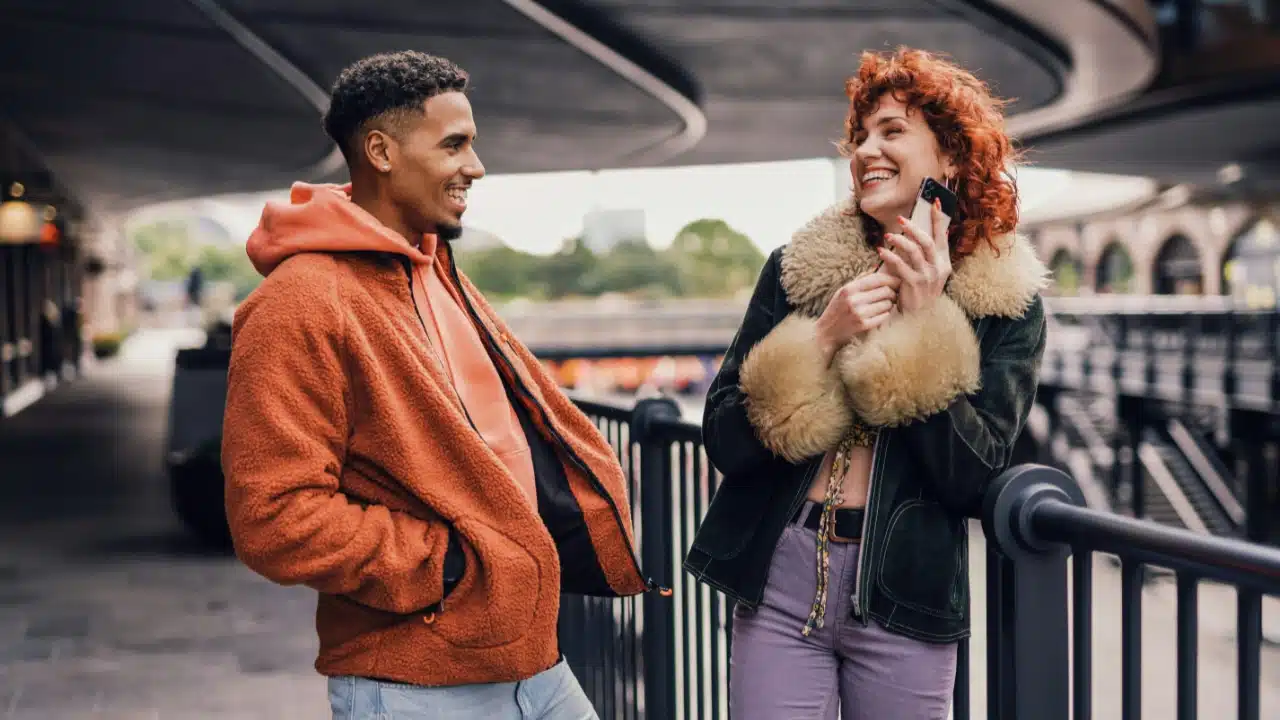 A man and woman in warm jackets laugh while standing together on an outdoor walkway.