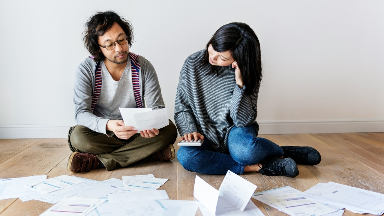 A man and woman sit cross-legged on a floor surrounded by scattered papers and envelopes.