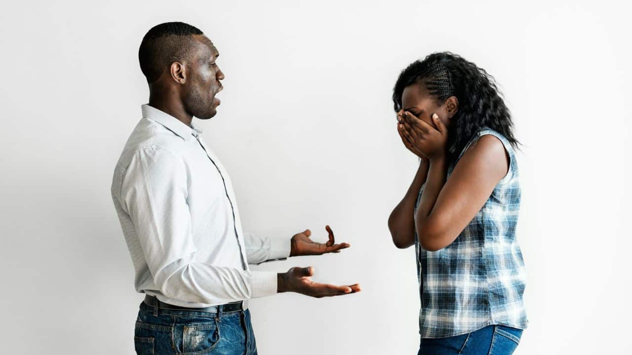 A man in a white shirt gestures toward a woman who is covering her face with hands.