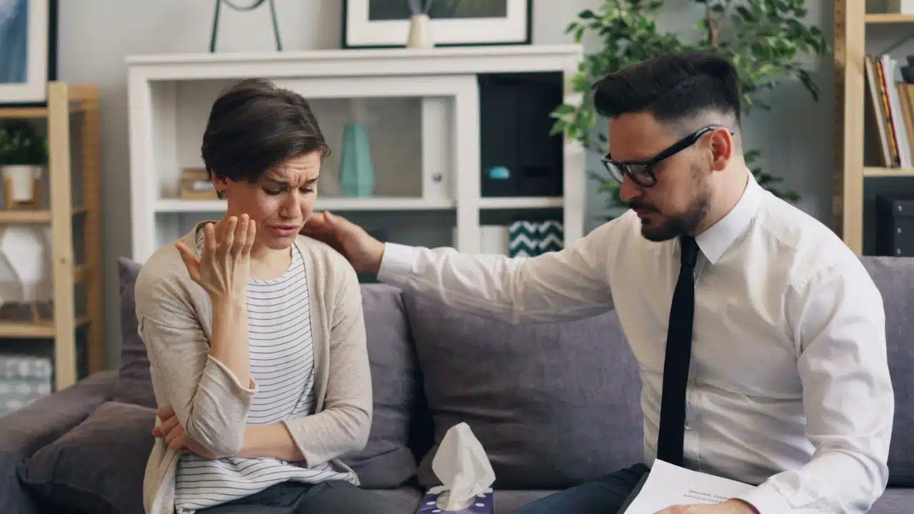 A woman crying on a couch while a man in a tie places a hand on her shoulder.