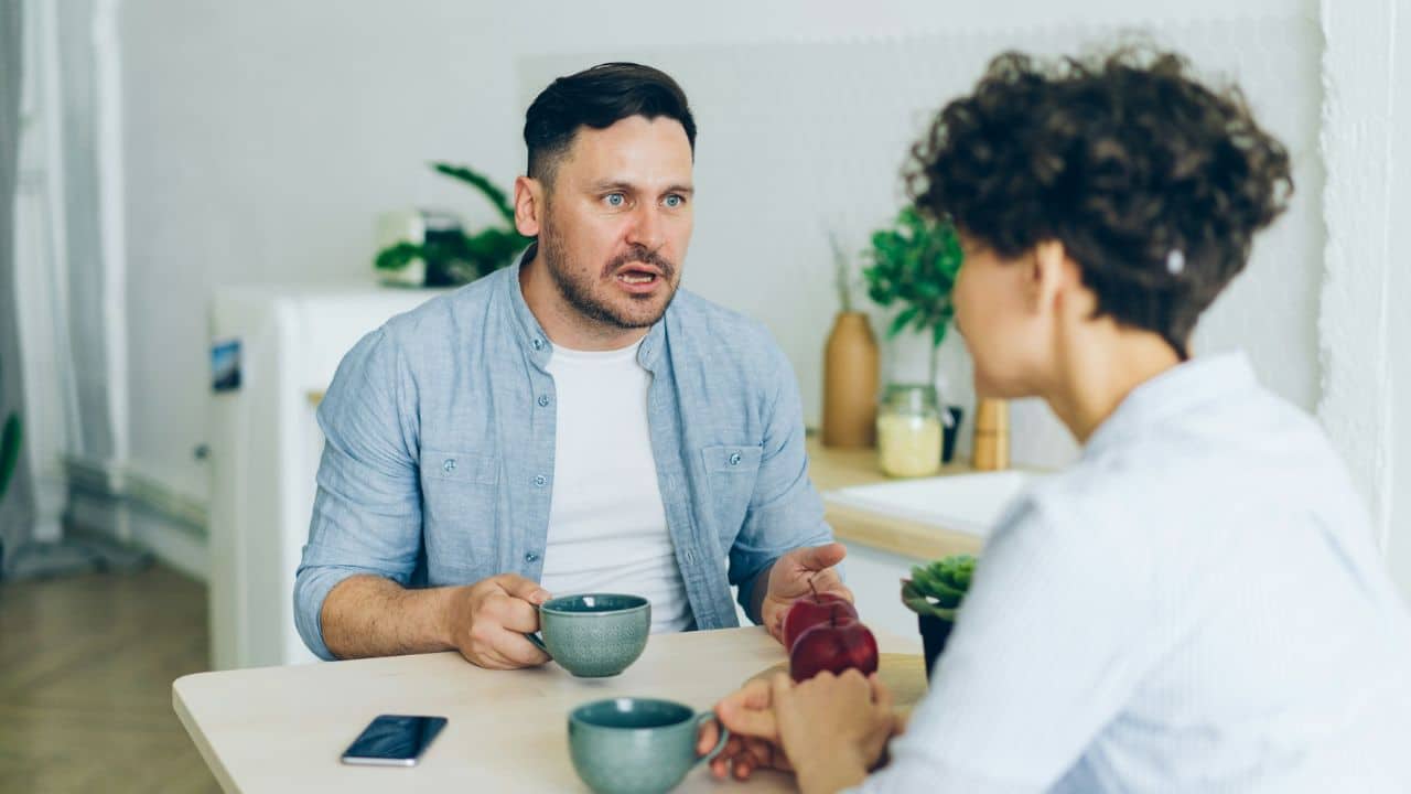 A man gestures while speaking to a woman across a table with two mugs.