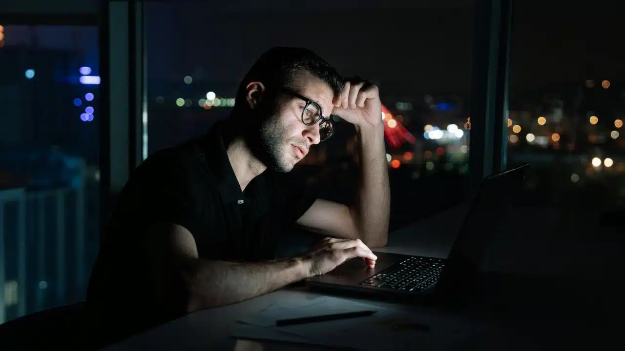 A man wearing glasses works on a laptop at night with city lights visible behind him.