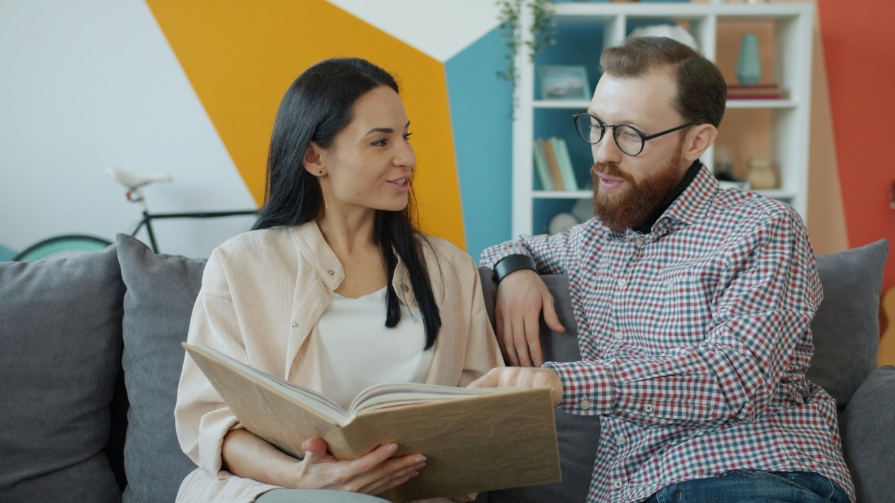 A woman and a man with glasses sit on a sofa looking through a large book.