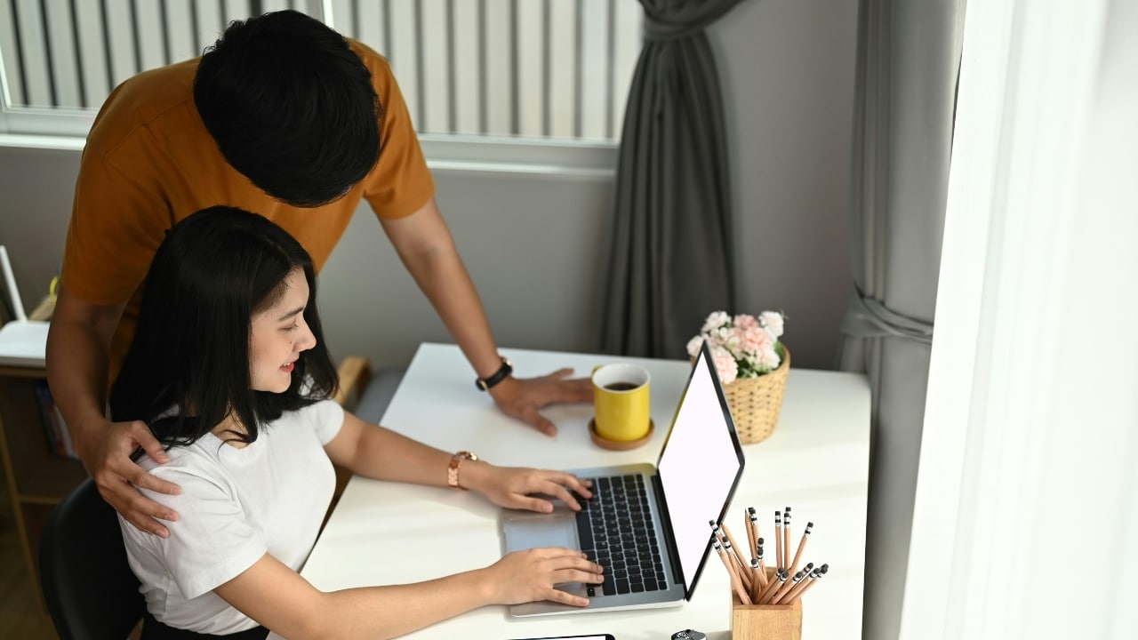 A woman types on a laptop while a man stands behind her with a hand on her shoulder.