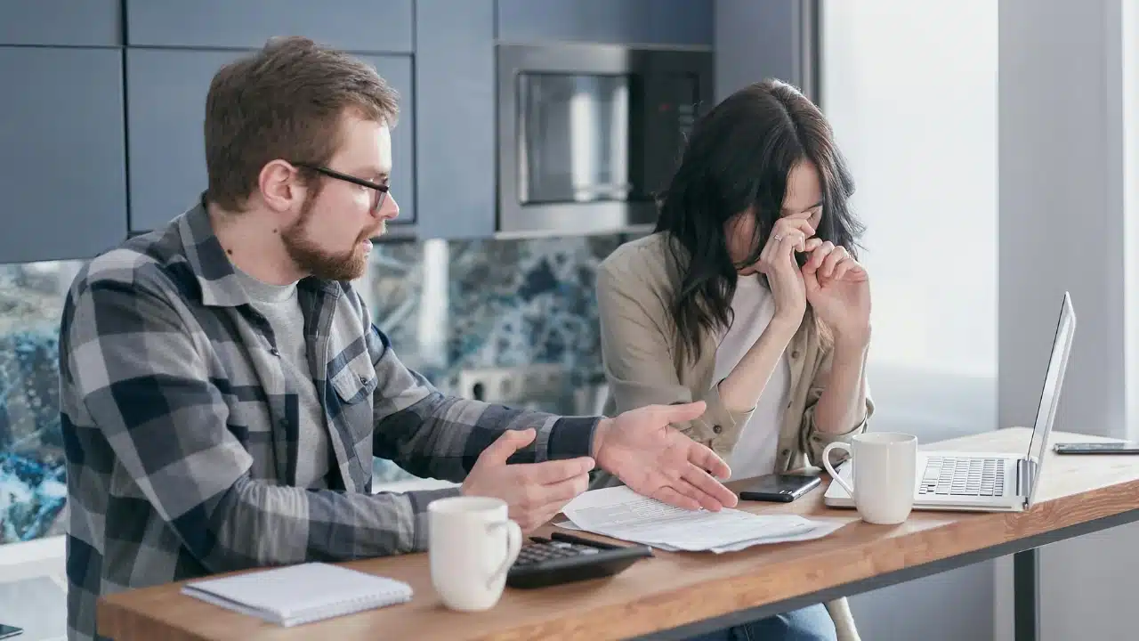 A man gestures toward papers and a calculator while a woman sits nearby with her head down.
