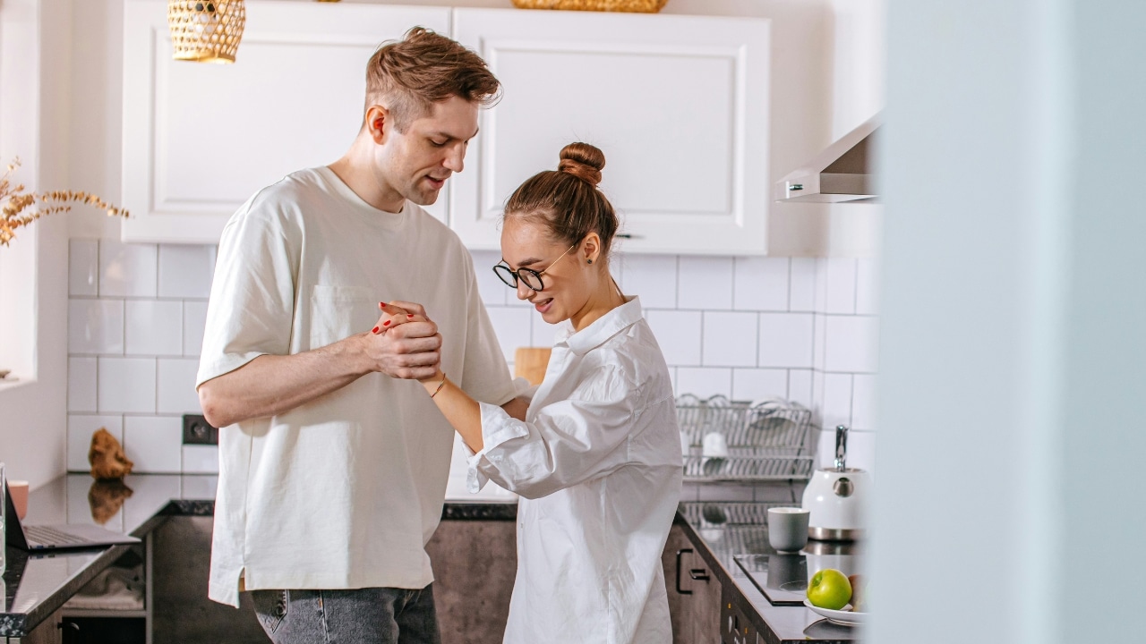 A man and woman hold hands and dance together in a bright, modern kitchen.