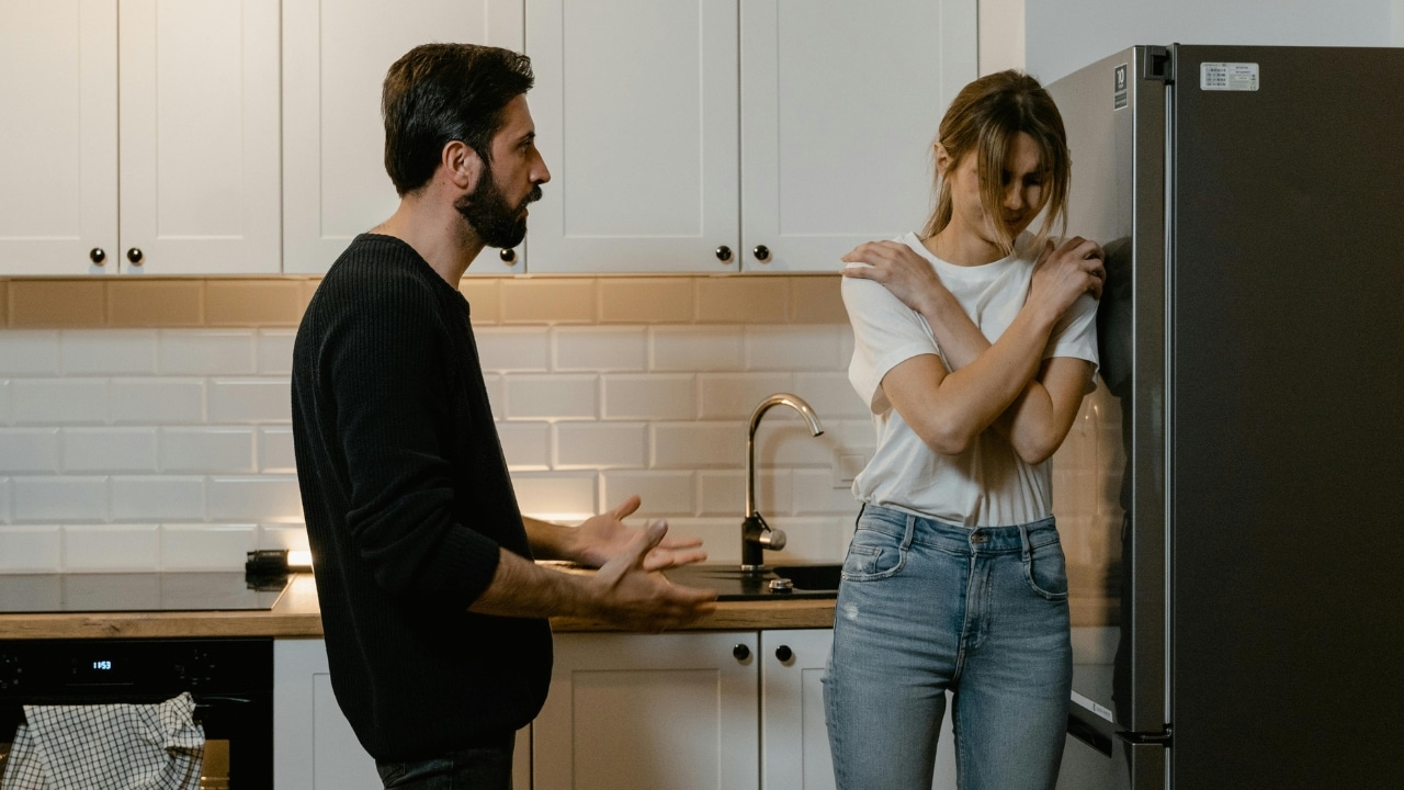 A man gestures toward a woman standing with her arms crossed in a white kitchen.