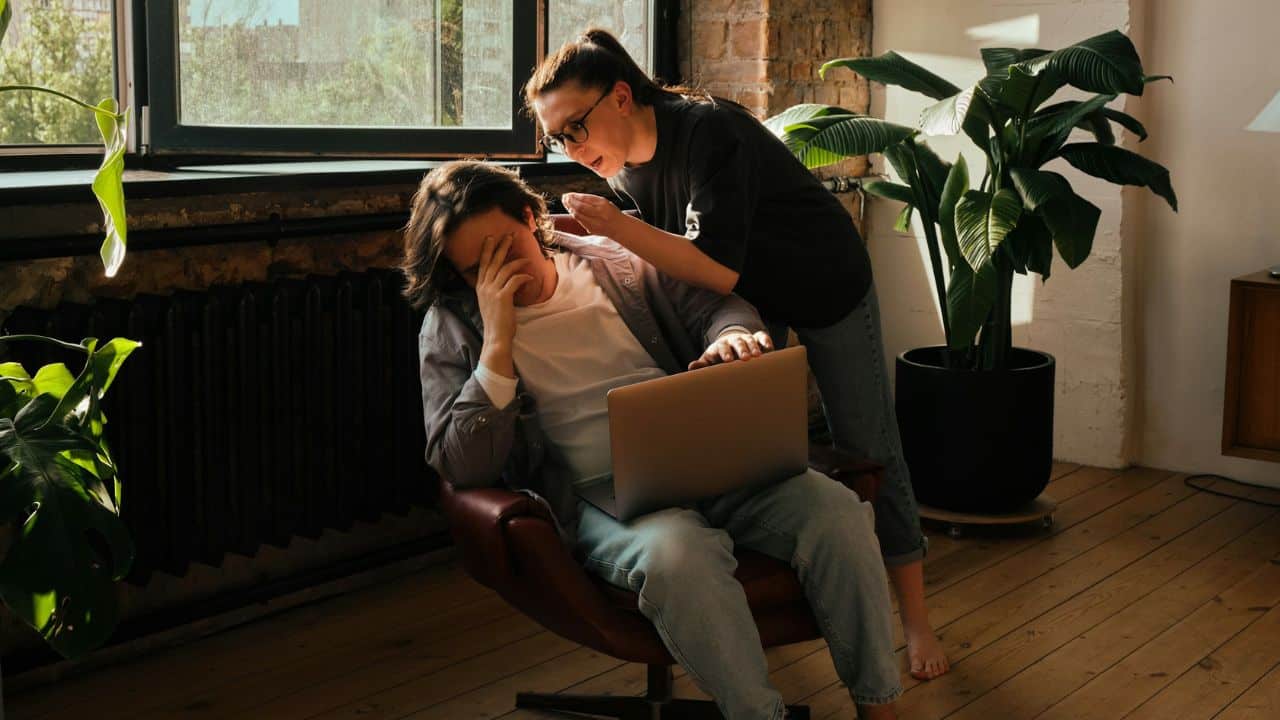 A woman stands behind a seated man who has his hand over his face.