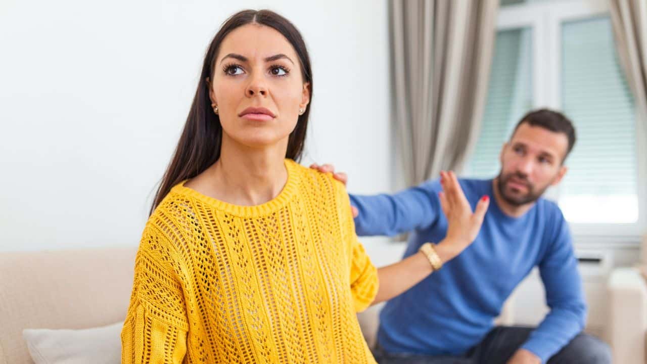 A woman looks away while a man behind her reaches out with his hand.