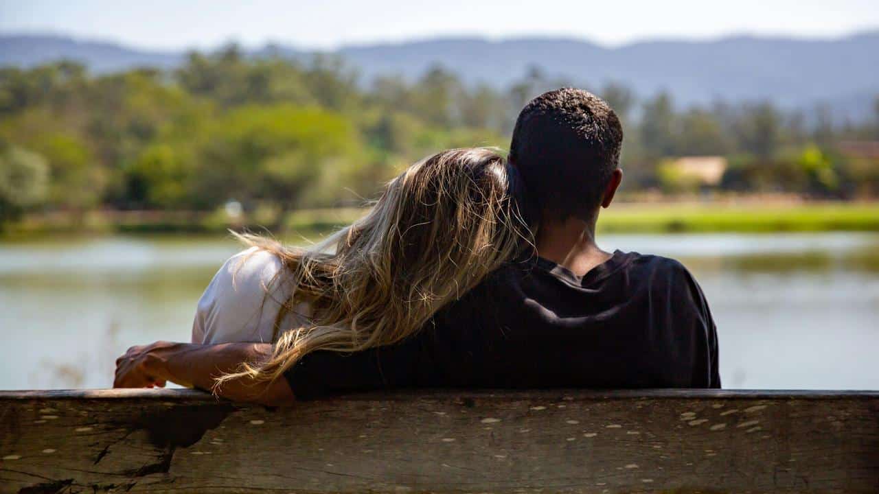 A couple sitting on a bench by the water with heads resting together.