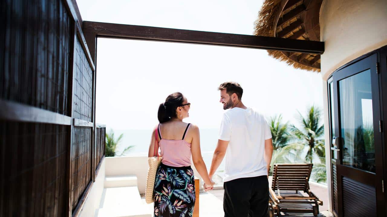 A couple holds hands while walking toward a sunny terrace with a sea view.