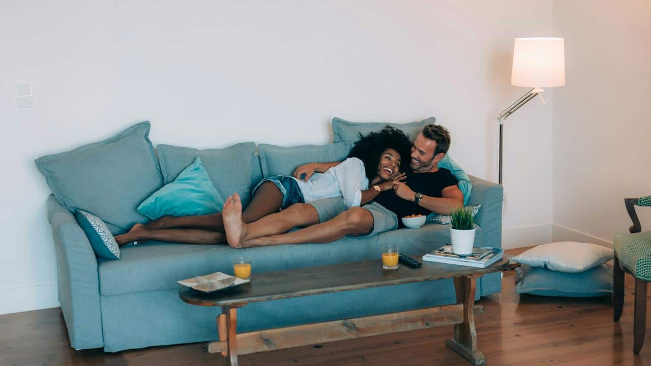 A couple relaxes together on a couch, laughing and enjoying snacks at home.
