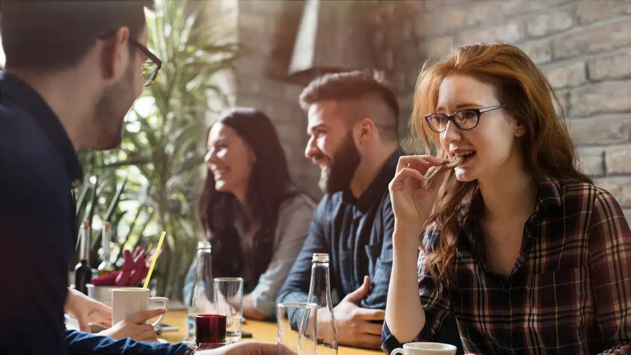 A woman in glasses eats a cracker at a table with three other people present.