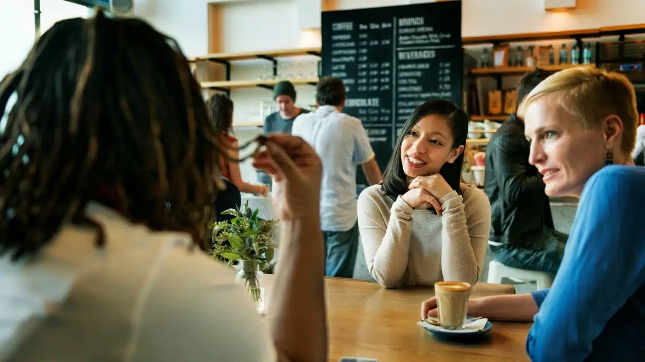 Two women sitting at a cafe table talking, with the back of another person's head visible.